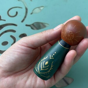 Puede incluir: Una mu&ntilde;eca de madera pintada a mano con cabeza marr&oacute;n y cuerpo verde. La mu&ntilde;eca tiene detalles pintados en oro en el cuerpo, incluyendo una flor en la cabeza y un dise&ntilde;o de hojas en el cuerpo.