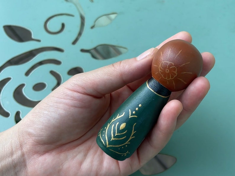 Puede incluir: Una mu&ntilde;eca de madera pintada a mano con cabeza marr&oacute;n y cuerpo verde. La mu&ntilde;eca tiene detalles pintados en oro en el cuerpo, incluyendo una flor en la cabeza y un dise&ntilde;o de hojas en el cuerpo.
