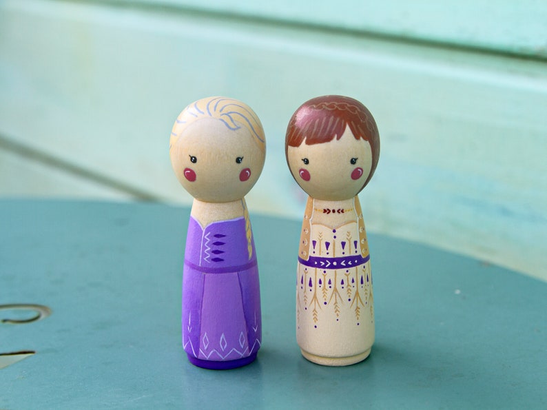 Puede incluir: Dos mu&ntilde;ecas de madera de clavija pintadas a mano vestidas con vestidos morados y beige con detalles intrincados. Las mu&ntilde;ecas tienen cabello rubio y casta&ntilde;o y caras pintadas con mejillas sonrosadas.