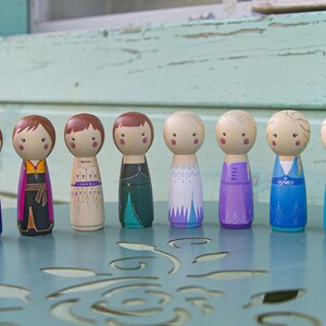 Puede incluir: Un conjunto de diez mu&ntilde;ecas de madera vestidas con atuendos coloridos. Las mu&ntilde;ecas llevan todas vestidos diferentes y tienen diferentes peinados. Las mu&ntilde;ecas est&aacute;n de pie sobre una mesa con un patr&oacute;n decorativo.