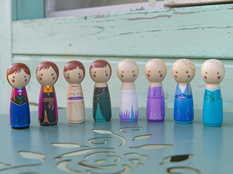 Puede incluir: Un conjunto de diez mu&ntilde;ecas de madera vestidas con atuendos coloridos. Las mu&ntilde;ecas llevan todas vestidos diferentes y tienen diferentes peinados. Las mu&ntilde;ecas est&aacute;n de pie sobre una mesa con un patr&oacute;n decorativo.