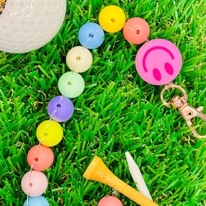 Puede incluir: Un porta bolas de golf con cuentas de colores arcoíris con un amuleto de cara sonriente rosa. El porta bolas está sobre una superficie de césped artificial verde con un tee de golf y una bola de golf blanca.