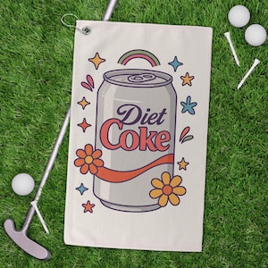 May include: Golf towel with a Diet Coke can design, featuring a rainbow, stars, and flowers. The towel is cream-colored and lies on green grass next to a golf club, golf balls, and tees.
