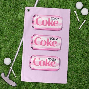 May include: A pink golf towel featuring three Diet Coke can illustrations. The towel is next to a golf club, two golf balls, and two tees, all on a green grass background.