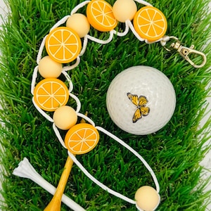 Puede incluir: Una bola de golf blanca con un diseño de mariposa, un porta bolas de golf blanco y naranja con rodajas de naranja, un tee de golf blanco y un tee de golf de madera sobre una superficie de hierba verde.