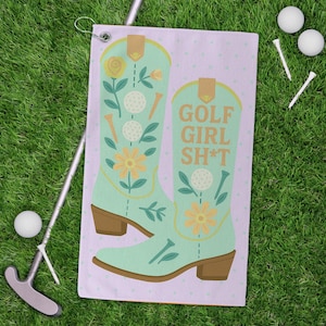 Puede incluir: Una toalla de golf con un diseño de bota de vaquero verde claro, con detalles florales y de pelota de golf. La toalla tiene el texto "GOLF GIRL SH*T". Un putter, pelotas de golf y tees están sobre la hierba verde.