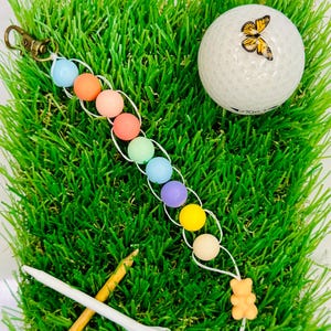 May include: A golf ball with a butterfly design, a golf tee, and a colorful beaded keychain with a gummy bear charm. The items are on a green artificial grass surface.