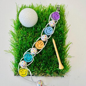 May include: A white golf ball, a golf tee, and a white bracelet with colorful smiley face charms and sprinkles on a green artificial grass surface.