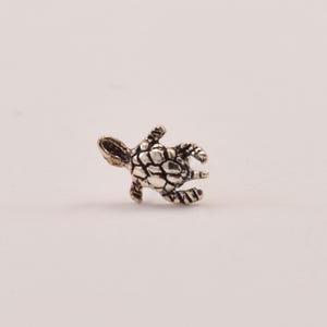 May include: A small, detailed silver-colored turtle figurine. The turtle has a textured shell and visible flippers and head. The figurine is likely a charm or pendant, suitable for jewelry or craft projects.