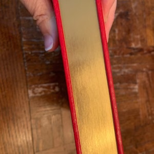 May include: The spine of a red hardback book with a gold-coloured cover. The book has a decorative white and red pattern along the top edge.