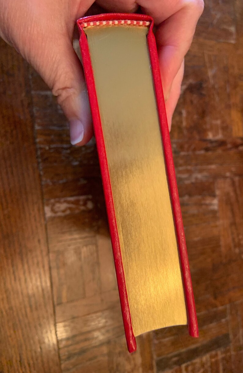 May include: The spine of a red hardback book with a gold-coloured cover. The book has a decorative white and red pattern along the top edge.