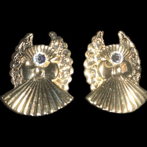 May include: A pair of gold tone earrings with a detailed design featuring a small clear rhinestone in the center of each earring.