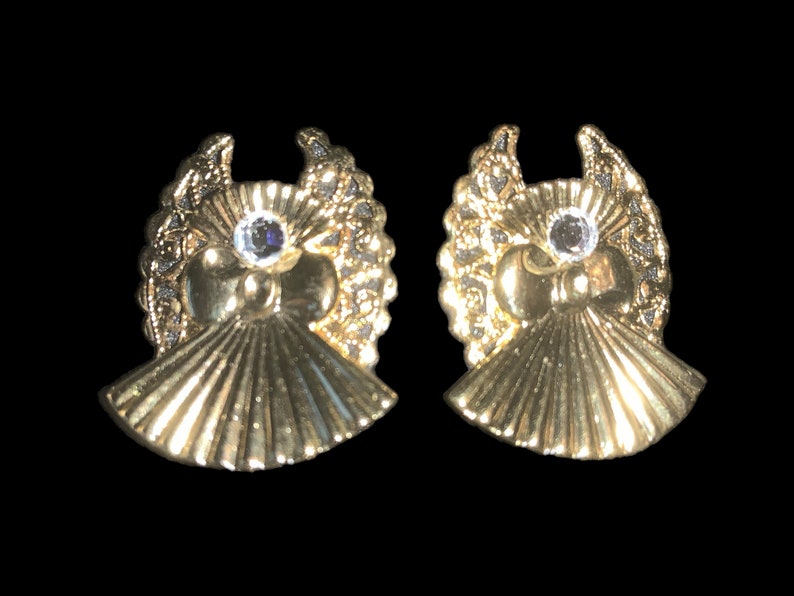 May include: A pair of gold tone earrings with a detailed design featuring a small clear rhinestone in the center of each earring.