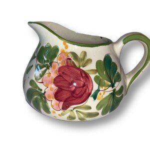 May include: A white ceramic creamer with a green and white floral design. The creamer has a rounded body and a handle. The floral design features pink, red, and yellow flowers.