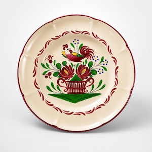 May include: A cream-colored ceramic plate with a scalloped edge and a dark red rim. The center features a hand-painted design of a rooster perched above a basket of red flowers and green leaves, surrounded by a decorative red leaf border pattern.