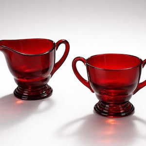 Peut inclure: Un ensemble de crémier et sucrier en verre rouge. Le crémier a un bec et une anse, tandis que le sucrier a deux anses. Les deux pièces ont un socle et sont en verre rouge transparent.