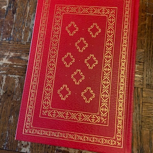 May include: A red hardback book with a gold embossed design on the cover. The design features a repeating pattern of stylised flowers and leaves.