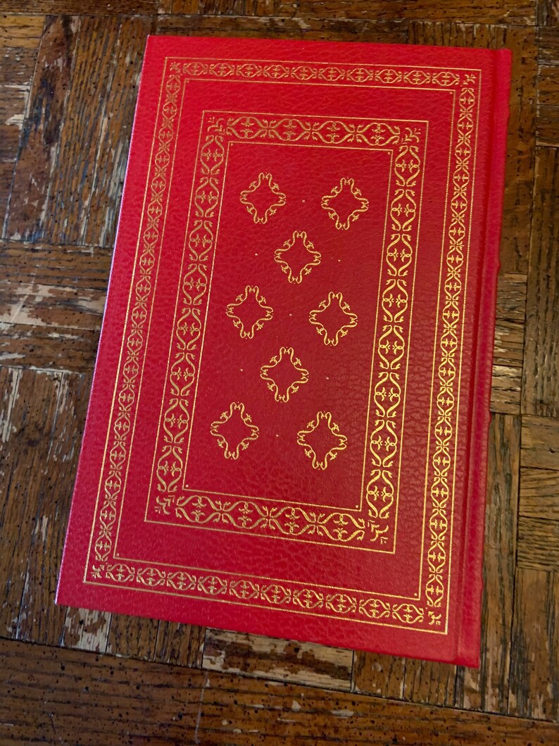 May include: A red hardback book with a gold embossed design on the cover. The design features a repeating pattern of stylised flowers and leaves.