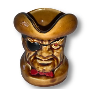 May include: A brown ceramic mug shaped like a pirate's head with a black eyepatch, brown hat, and red bow tie.