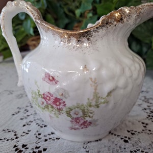 May include: White porcelain pitcher with a floral design and gold accents. The pitcher has a curved handle and a spout.