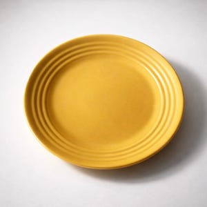 May include: A round, mustard-yellow ceramic plate with concentric ridged detailing around the rim. The plate is centered on a white background, casting a soft shadow. This dishware is suitable for serving meals.