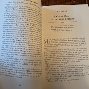 May include: An open book with a cream coloured cover and black text. The book is open to a chapter titled "A Great Heart and a Small Fortune". The text on the page is a quote from Don Juan, Canto 1, Stanza 75. The book is resting on a wooden surface.