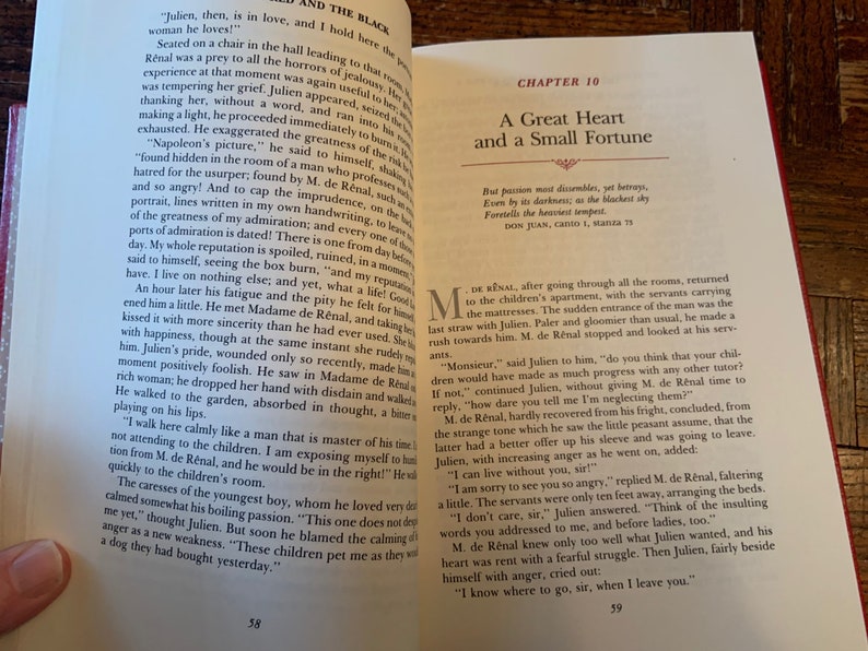 May include: An open book with a cream coloured cover and black text. The book is open to a chapter titled "A Great Heart and a Small Fortune". The text on the page is a quote from Don Juan, Canto 1, Stanza 75. The book is resting on a wooden surface.