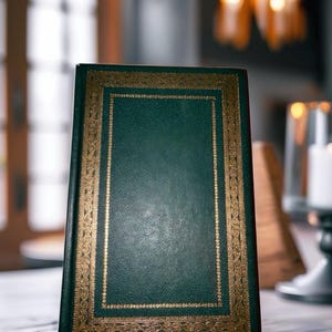 May include: A hardcover book with a dark green cover and gold-colored decorative border. The book is standing upright on a wooden surface. The background is blurred, suggesting a room setting with soft lighting.