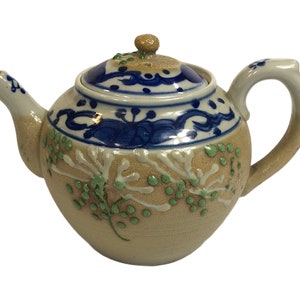May include: A small, cream-colored teapot with a blue and white floral design. The teapot has a lid and a handle.