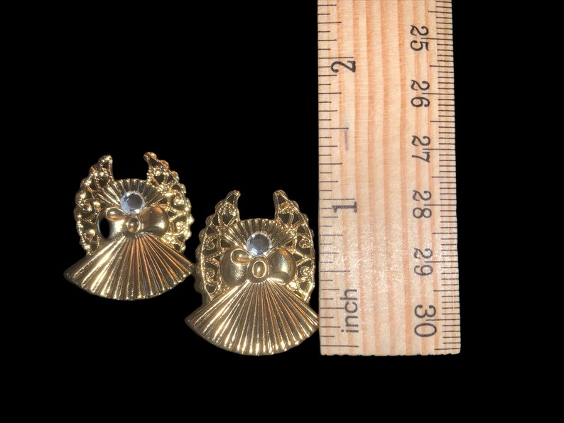 May include: Two gold angel-shaped ornaments with a clear rhinestone in the center of each. The ornaments are approximately 1 inch in diameter.