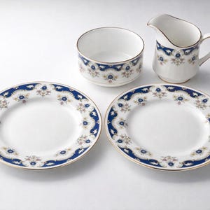 Könnte beinhalten: Ein weißes Porzellan-Teeservice mit blauem Blumenmuster. Das Set beinhaltet zwei Teller, eine Zuckerdose und einen Sahnegießer. Die Teller haben einen goldenen Rand. Der Sahnegießer hat einen Griff und einen Ausguss.
