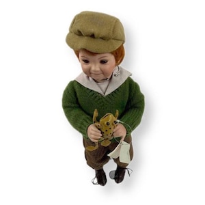 May include: A porcelain doll boy wearing a green sweater, brown trousers, and a brown cap. He is holding a small, brown frog in his hands.