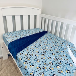 May include: A white cot with a light blue fitted sheet patterned with a cartoon dog design. A dark blue blanket is placed over the sheet. The background is a plain white wall.
