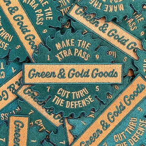 May include: A close-up of several embroidered patches with a green and gold color scheme. The patches feature different designs, including "Make the Xtra Pass", "Cut Thru the Defense", and "Green & Gold Goods".