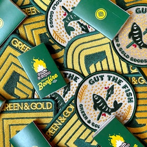 May include: A collection of green and gold embroidered patches with various designs, including a fish, a bonfire, and a campfire. The patches are arranged on a table with a green and gold background.
