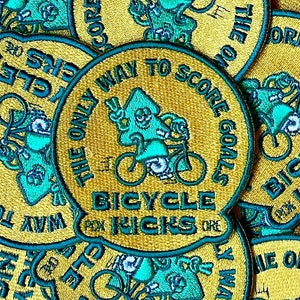 May include: Embroidered patches with a yellow background and teal accents. The patches feature a bicycle and the text "THE ONLY WAY TO SCORE GOALS BICYCLE KICKS". The patches have a circular shape.