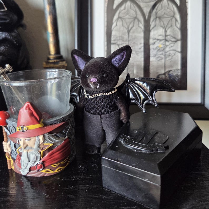 Puede incluir: Un peluche de murci&eacute;lago negro con detalles morados, que lleva un collar de cadena y pantalones oscuros, se encuentra cerca de una caja negra con forma de ata&uacute;d. Tambi&eacute;n se puede ver un candelabro con tem&aacute;tica de mago y una obra de arte enmarcada de ventana g&oacute;tica.