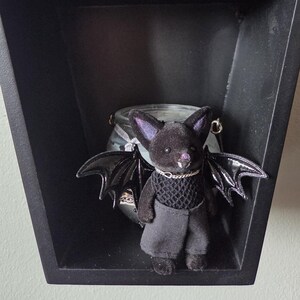 Puede incluir: Una figura de murci&eacute;lago de peluche negro con alas negras brillantes, que lleva una camiseta de malla negra y pantalones grises. El murci&eacute;lago tiene orejas forradas de p&uacute;rpura y un collar de cadena plateada. Se exhibe en un estante negro y geom&eacute;trico.