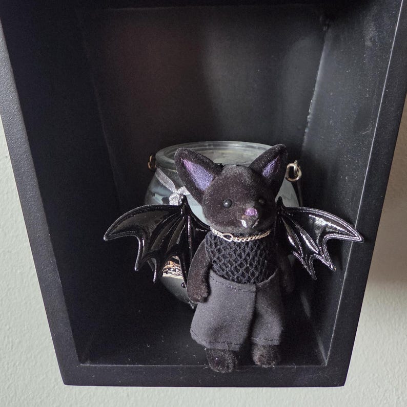 Puede incluir: Una figura de murci&eacute;lago de peluche negro con alas negras brillantes, que lleva una camiseta de malla negra y pantalones grises. El murci&eacute;lago tiene orejas forradas de p&uacute;rpura y un collar de cadena plateada. Se exhibe en un estante negro y geom&eacute;trico.