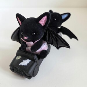 May include: Two black bat figurines with pink and blue accents. One bat is seated in a black toy car with a moon and bat graphic. The bats have large ears and glossy black wings. The bat in the car is wearing a pink outfit.
