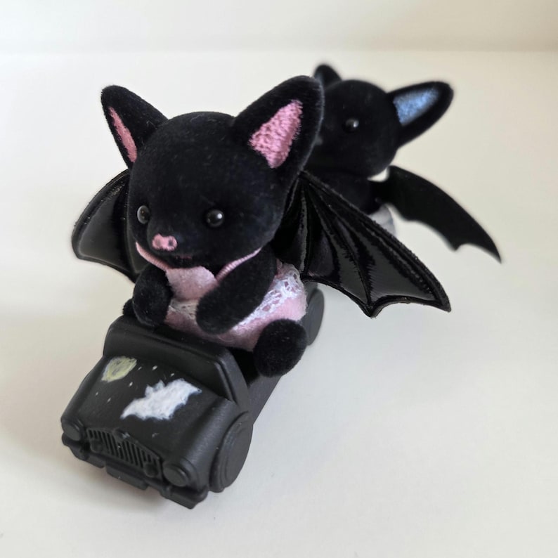 May include: Two black bat figurines with pink and blue accents. One bat is seated in a black toy car with a moon and bat graphic. The bats have large ears and glossy black wings. The bat in the car is wearing a pink outfit.