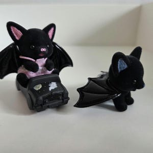 May include: Two black bat figurines. One bat is seated in a black toy car with a painted night sky design. The other bat is standing, with its wings spread. Both bats have pink inner ears and are made of a soft, plush material.
