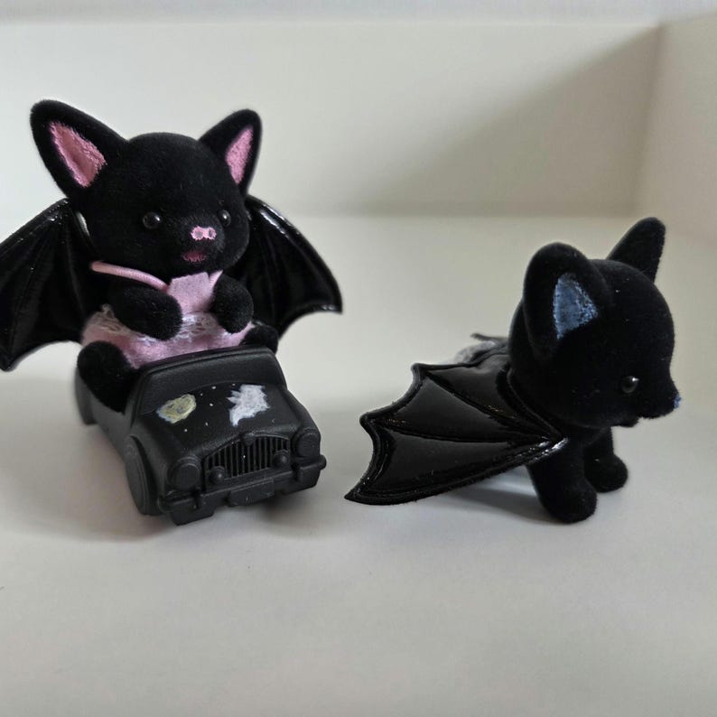 May include: Two black bat figurines. One bat is seated in a black toy car with a painted night sky design. The other bat is standing, with its wings spread. Both bats have pink inner ears and are made of a soft, plush material.