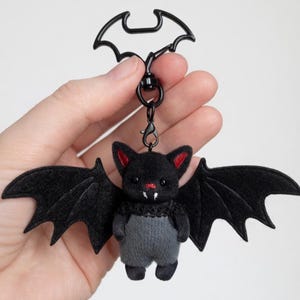 May include: A plush black bat keychain with red ear accents and a small red nose. The bat has outstretched felt wings and a gray body. The keychain features a black bat-shaped clip.