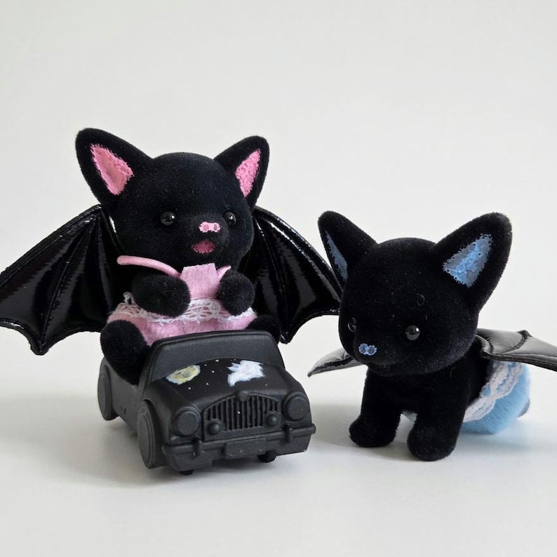 May include: Two black bat figurines with pink and blue accents. One bat is seated in a black toy car with a moon and star design. The other bat is standing. Both have black wings.