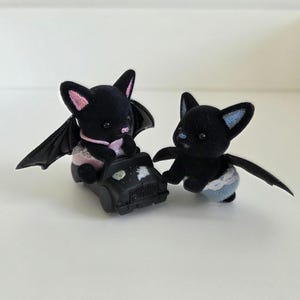 May include: Two black bat figurines with pink and blue accents. One bat is seated in a black toy car with a painted night sky design. The other bat stands nearby. Both have wings and wear diapers.