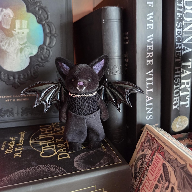 Puede incluir: Una mu&ntilde;eca de murci&eacute;lago de peluche con pelaje negro, orejas moradas y alas negras brillantes. Lleva un peque&ntilde;o collar de cadena plateada y pantalones grises oscuros. La mu&ntilde;eca est&aacute; posada sobre una pila de libros oscuros, incluido uno titulado "CTHUTHU DARK ARTS."