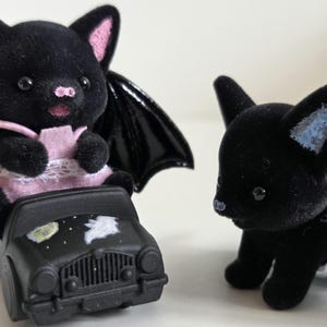 May include: Two black bat figurines, one in a pink dress with black wings, seated in a black car with a night sky design. The other bat figurine is standing nearby. The bats have pink ears and noses.