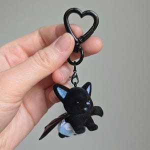 May include: A black keychain featuring a plush bat with blue accents on the ears and a small blue cloud on the rear. The keychain has a black heart-shaped clasp.