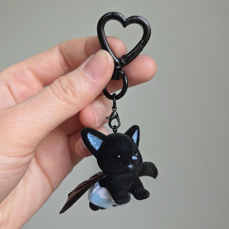 May include: A black keychain featuring a plush bat with blue accents on the ears and a small blue cloud on the rear. The keychain has a black heart-shaped clasp.